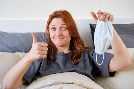 The recovered woman took off her mask and shows a thumbs up. Girl having had coronavirus at home in bedの写真素材