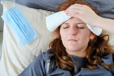 Woman measures temperature while lying in bed with a compress on her foreheadの写真素材