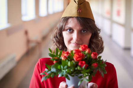 A young woman in a military uniform of the Soviet Union holding a bouquet of red roses. A girl in a red dress with flowers stands in a long corridor. Russia, Moscoの写真素材