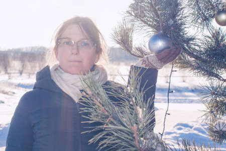 Young woman stands on the background of a winter landscape and holds a Christmas tree toyの写真素材