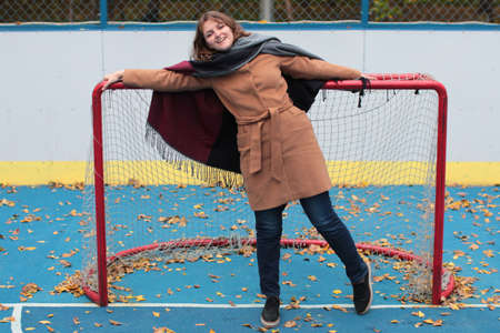 Outdoor play area on the street. A young woman stands at the gate of the hockey field. Autumn weather on the open sports field with blue coating.の写真素材