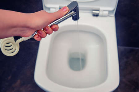 Hygienic shower in hand with a stream of water, close-up on a white toilet bowlの写真素材