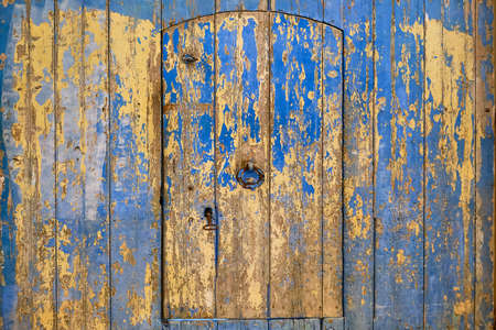 Old wooden door covered with peeling blue paint. A yellow wooden wall and a semicircular Arabic door with a knocker handle.の写真素材