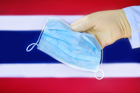 Medical mask in the hands of a doctor and the Thailand flag, copy space. Protective glove with mask close-up on the background of the state Thai symbolの写真素材