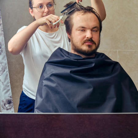 Wife cuts a man's hair with scissors in front of the mirror, copy space. The concept of unkempt appearance and problems in isolation from the coronavirus pandemicの写真素材