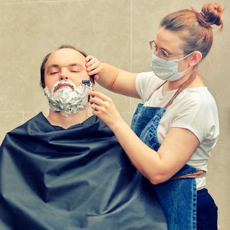 Woman barber in a medical mask shaves a man with a safety razor. Concept of unkempt appearance and problems in isolation from the coronavirus pandemicの写真素材