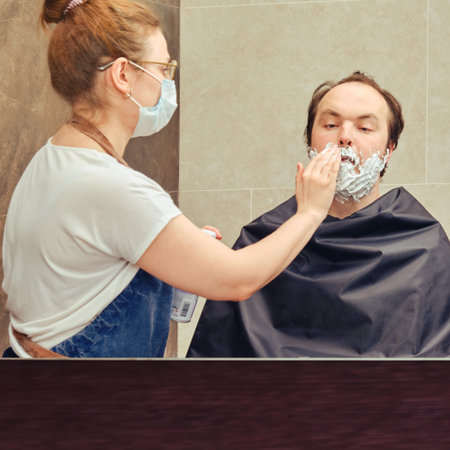 Barber in a medical mask applies shaving foam to a man in a home bathroom, reflected mirror, copy...