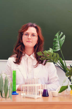 Cute teacher in a white coat sits at a desk. Scientific experiments in a school class. Experiments with chemical reagents at the school deskの写真素材