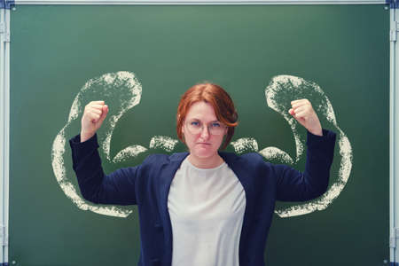 School teacher raised her hands to show strength at the blackboard. A female teacher with muscles drawn in chalkの写真素材