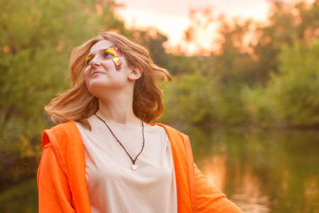 Summer portrait of a girl on a pond background. Woman with face painting on the background of the lake. Developing hair in the rays of the setting sun.の写真素材