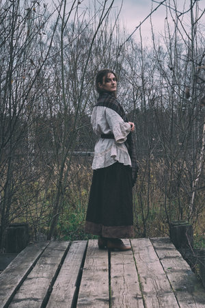 A woman in a headscarf looks from the bridge at the old pond. Girl in a retro dress on a platform in an autumn gloomy landscapeの写真素材