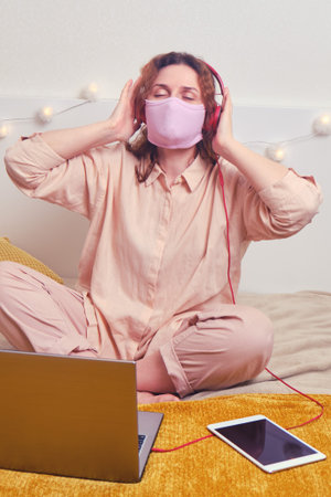 Young girl in the face mask dances sitting on the home bed. Happy woman in medical mask is resting...