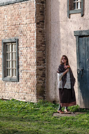 A woman with flowers in retro clothes stands at the door of an old houseの写真素材