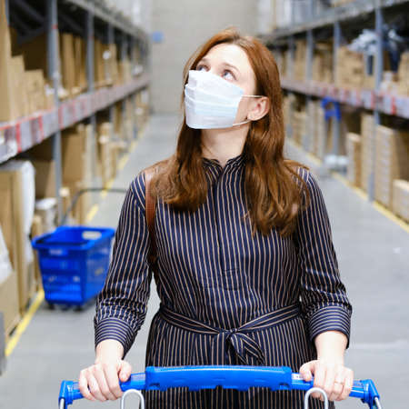 Woman in black clothes and a medical mask with a shopping cart in a storehouse during quarantine of coronavirusの写真素材