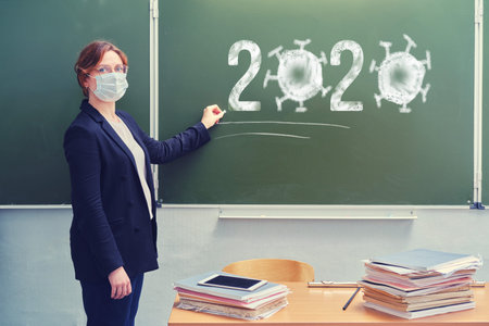 A teacher is standing in a medical mask at the blackboard with chalk 2020, copy space. Woman school...