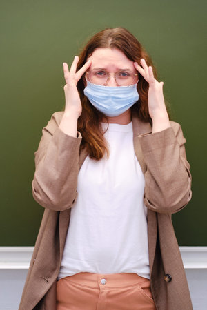 Teacher in a face mask stands with a headache. Concept of problems at school during the coronavirus epidemicの写真素材