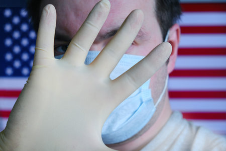 A man in a medical mask covers his hand against the background of the US flag