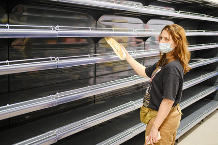 A young woman in a medical mask takes the last loaf of bread from the empty shelves of the store