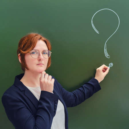 School teacher with a sad face writes in chalk a question mark on a blackboard. Upset woman teacher on green backgroundの写真素材