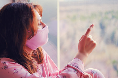 A young woman in a medical protective mask is sad at the window because of the coronavirus epidemic