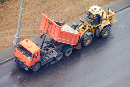 Asphalt is poured from the truck body into the bucket of a bulldozer during road works on a city...