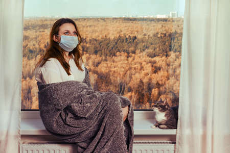 A young woman sits on a windowsill in a medical mask in quarantine. Concept of coronavirus quarantine and home isolationの写真素材