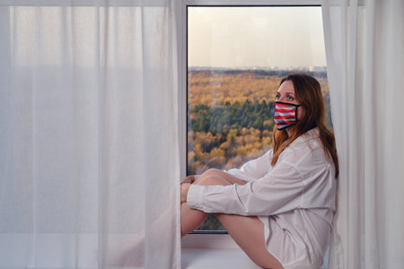 Woman in medical mask with usa flag sits on windowsill by window. Concept of coronavirus quarantine...