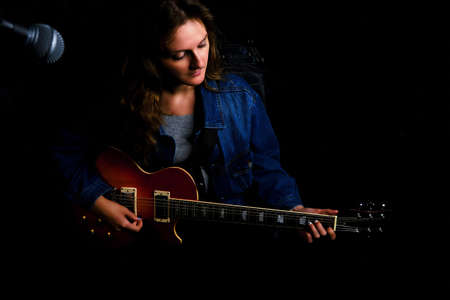 A woman with a red electric guitar records music at a recording studio, copy space. Female guitarist sitting behind a microphone on a black background.の写真素材