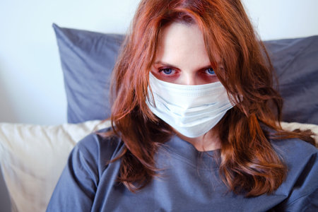 Portrait of a sick woman in a mask on the bed, close-up. Face of a woman with red hair in a medical...