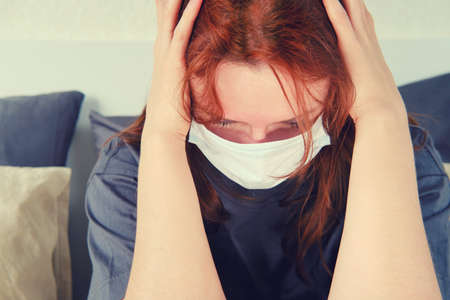 A woman in a medical mask has a headache. woman with a sore head sits on the bedの写真素材
