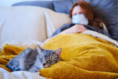 Redhead woman in medical mask sick with coronavirus and cat are lying on the bed.の写真素材