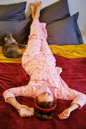 Red-haired woman in red pajamas on a red bed at home next to pet. A woman listens to music on headphones with a cat during a coronavirus epidemicの写真素材