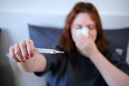 A sick woman with a runny nose holds out a thermometer at arm's length