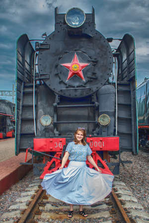 woman with billowing dress against vintage background of locomotive. Retro train of USSR with a red star. Young woman in a white dress on the rails at steam locomotive.の写真素材