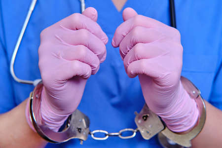 A doctor in a blue uniform with handcuffs on his hands, close-up. Medic in shackles, the concept of coronavirus quarantine in the worldの写真素材