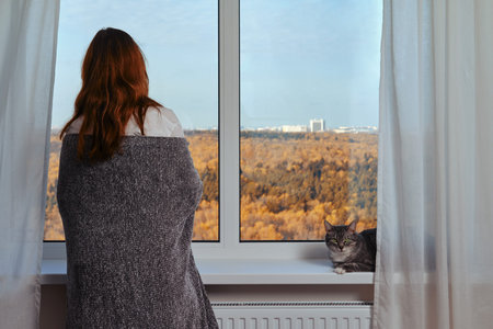 Adult woman at the window wrapped in a gray blanket next to a cat. Autumn nature outside the window...