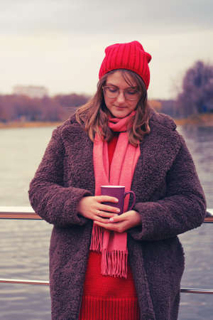 Young woman in coat with a cup near the autumn pondの写真素材