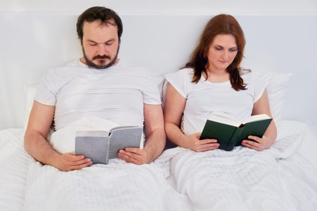 A man and a woman read books lying in a white bed, lifestyle. Husband and wife at home under...