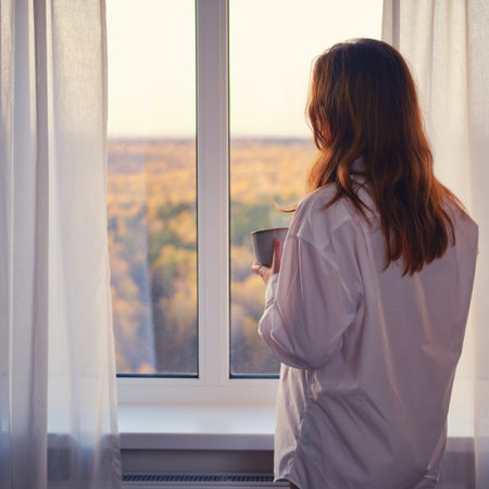 A woman stands by the window over the autumn forest. Young woman with a cup in her hands looks at...