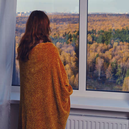 A red haired woman stands wrapped in a blanket at the window and looks out at the autumn forest. Sad autumn weather and home comfortの写真素材