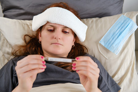 Woman with red hair lies in bed sick with coronavirus with a cold compress on her headの写真素材