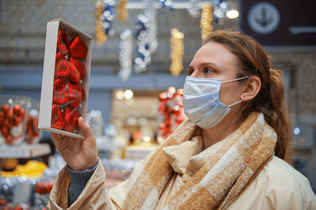 A woman in a face mask with a heart shaped toy in her hand buys Christmas decorations. Lifestyle,...