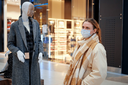 Woman in mask face next to clothing mannequin in shopping mall. Living with the coronavirus pandemic