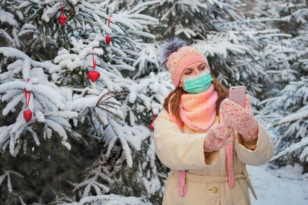 Woman in face mask communicates online chatting for the new year. Internet by phone in the winter park near the Christmas treeの写真素材