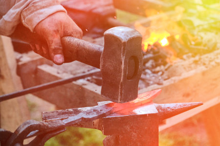 Blacksmith hammer hits the hot metal. Forging of iron objects in a retro forge. The process of working on the manufacture of weapons blacksmith on the anvil.の写真素材