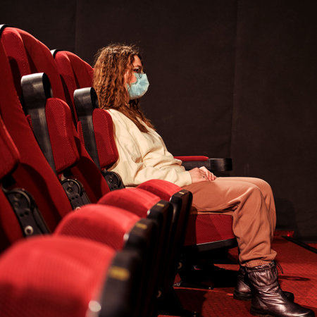 Adult woman in a medical mask watches a movie in an empty theater hall. Bankruptcy of entertainment companies during the flu virus epidemicの写真素材