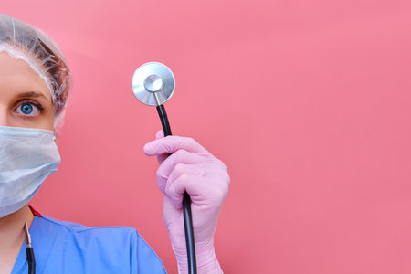 Doctor with a stethoscope in hand on a pink background, copy space for text. Woman nurse in a medical mask helps patients with coronavirus.の写真素材