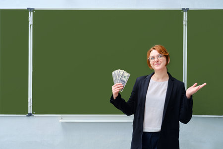 Happy teacher holding money, us dollars at the blackboard, copy spaceの写真素材