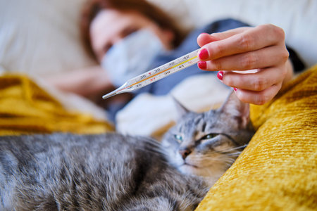 A young redhead woman looks at a thermometer next to a cat. Sick woman lies at home in a medical...