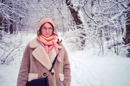 A woman in a white fur coat and pink hat stands in a snow-covered winter forest. Portrait of an adult woman with glasses and a bag among the trees in the park.の写真素材
